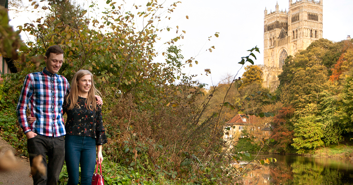 man and woman walking past the River Wear with Durham Cathedral in background.